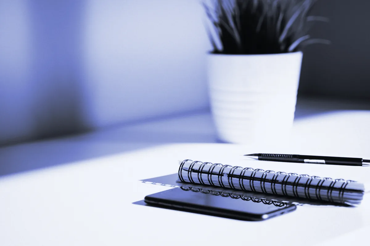 Cuaderno de espiral, bolígrafo y móvil sobre una mesa blanca junto a una planta